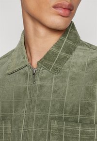 Olijfgroen corduroy shirt met een ritskraag, voorzien van een ruitpatroon en een borstzak. De textuur is ribbelig en zacht.