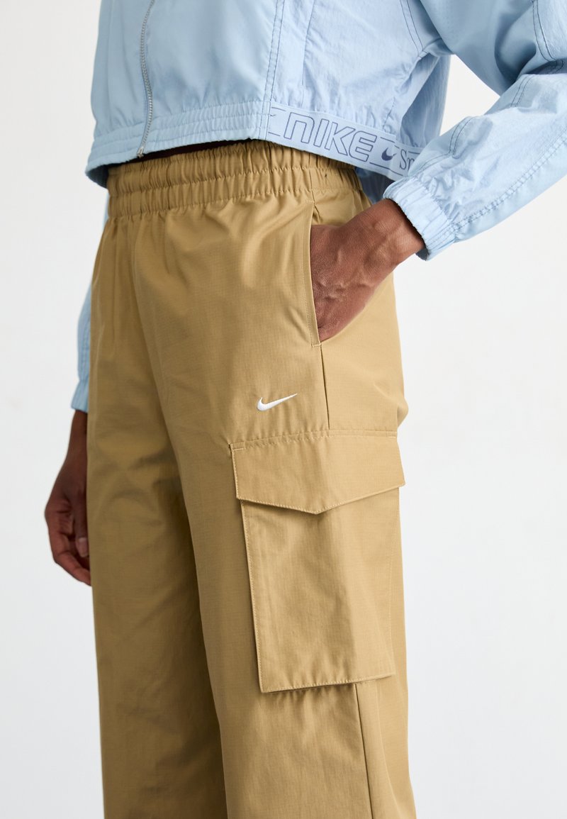 Pantaloni cargo Nike in tessuto beige, con cintura elastica, tasca laterale e logo Nike bianco sulla coscia sinistra.