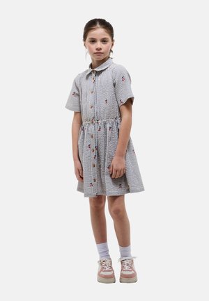 Jeune fille debout, portant une robe grise avec de petits motifs floraux rouges et noirs, des chaussettes blanches et des baskets roses.