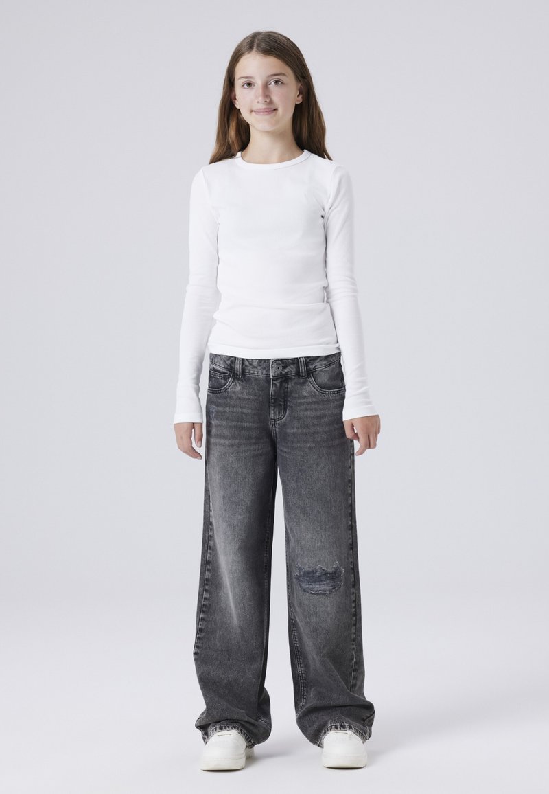 Bílá košile s dlouhým rukávem v kombinaci s volnými šedými džíny s vysokým pasem, které mají prvky opotřebení. Model má na sobě bílé tenisky.