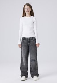 Camisa blanca de manga larga combinada con jeans grises de tiro alto y corte holgado que presentan detalles desgastados. La modelo lleva zapatillas blancas.