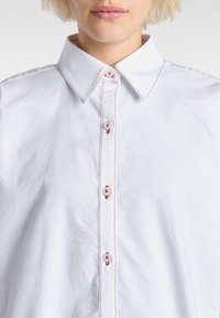 White collared shirt with red stitching and plain buttons, featuring a button-down front and a smooth, textured fabric.