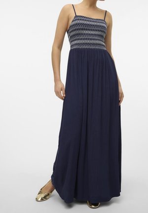 Kobieta w granatowej sukience maxi z marszczonym, wzorzystym górnym częścią i cienkimi ramiączkami, w połączeniu z błyszczącymi złotymi balerinami.