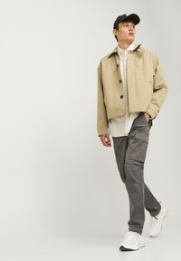 Beige kort jacka med kn�äppning, kombinerad med grå cargobyxor och vita sneakers. Modellen bär en svart keps och en vit skjorta under.