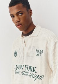 Jonge man draagt een crèmekleurige, gestructureerde trui met kraag, met erop geborduurd in donkergroen "New York Finale state league" en "MCM XCI".