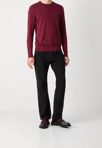Mann in einem burgunderfarbenen Langarmpullover, schwarzen Jeans und dunkelbraunen Slippern mit burgunderfarbenen Akzenten, der vor einem schlichten Hintergrund steht.