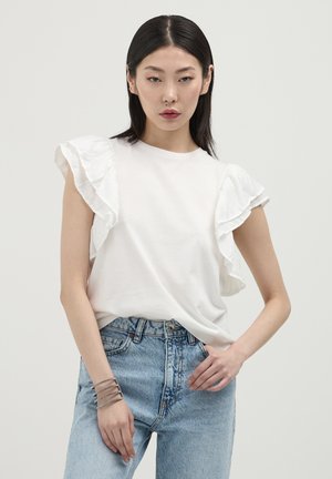 Haut blanc à manches courtes avec des détails de manches froncées. Associé à un jean en denim bleu clair taille haute. Les accessoires comprennent un bracelet avec plusieurs bandes.