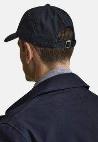 Marinblå basebollkeps med en tygrem och metallspänne på baksidan. Har en broderad logotyp nära justeringen. Bärs tillsammans med en jacka med krage.