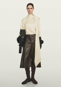 Crème lange mouw top met gedrapeerde sjaal, gecombineerd met hoog in de taille bruine leren culottes. Zwarte handschoenen en strakke bruine laarzen maken de look compleet.