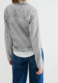 Šedá džínová bunda s přiléhavým střihem, se zadním dílem, manžetami na knoflíky a kontrastním prošíváním, v kombinaci s modrými džínami a bílou košilí.