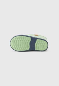 Suela de goma verde con un patrón ondulado, presentando un lado marino y un acento marrón claro. El zapato tiene una forma redondeada y una superficie de agarre texturizada.