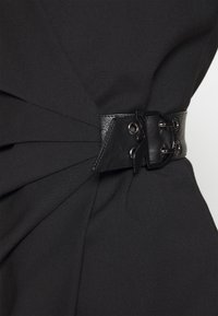 Schwarzer Stoffgürtel mit strukturiertem Finish, ausgestattet mit einer großen schwarzen Metallschnalle und Nieten, vor einem plissierten Kleidungsstück.