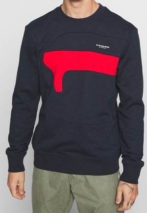 Osoba w granatowej bluzie z wyrazistym czerwonym geometrycznym paskiem na piersi oraz małym białym logo "G-Star Raw" na górze po prawej stronie.