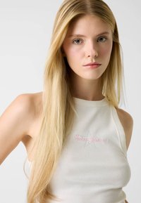 Witte geribbelde tanktop met roze en blauwe "Guess Jeans" borduursel. Heeft een hoge halslijn en mouwloos ontwerp. Recht, blond haar.