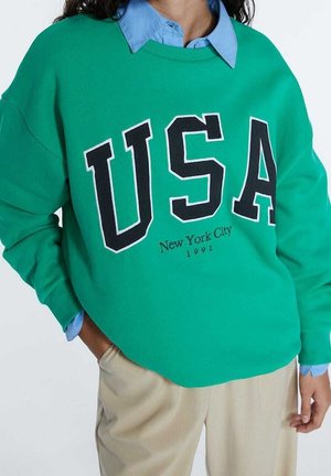 Personne portant un sweat-shirt vert avec "USA" et "New York City 1991" en texte noir sur une chemise bleue à col et un pantalon beige.
