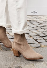 Cizme de gleznă bej din suede cu vârf ascuțit, detalii verticale cu rigle și un toc din lemn stratificat pe paviment de piatră cubică.