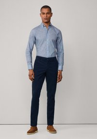 Camisa de botones azul claro con estampado, pantalones oscuros de color marino y zapatos de ante marrones. Diseño simple y ajustado con una textura suave y detalles sutiles.