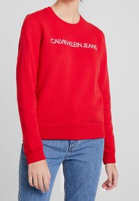 Sudadera roja de algodón, con escote redondo y puños acanalados. Lleva el texto blanco "CALVIN KLEIN JEANS" en la parte delantera.