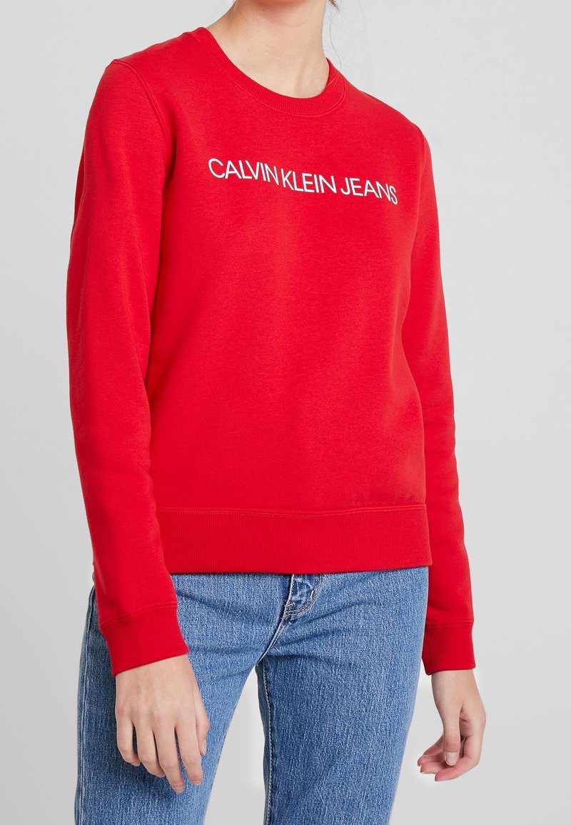 Sudadera roja de algodón, con escote redondo y puños acanalados. Lleva el texto blanco "CALVIN KLEIN JEANS" en la parte delantera.