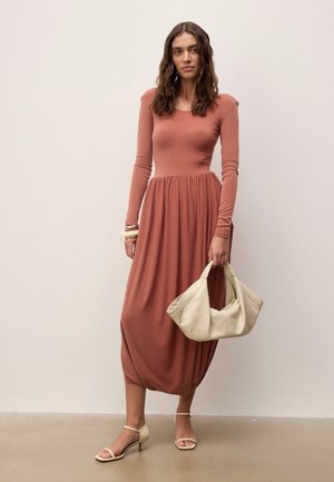 Femme en robe midi à manches longues rose poussière tenant un sac à main crème, portant des talons crème et des bracelets, debout devant un fond uni.