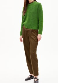 Frau in einem grünen Strickpullover, braunem Hemd mit Kragen, braunen plissierten Hosen und schwarzen Schnallenschuhen, die vor einem weißen Hintergrund steht.