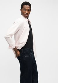 Νεαρός άνδρας με μαύρο πουκάμισο, ανοιχτό ροζ πουκάμισο με κουμπιά και σκούρο τζιν, στέκεται πλάγια με το χέρι στην πίσω τσέπη.