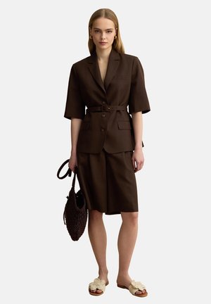 Mujer con blazer marrón oscuro con cinturón y pantalones cortos hasta la rodilla, sosteniendo un bolso tejido, calzando sandalias crema abiertas en los dedos.