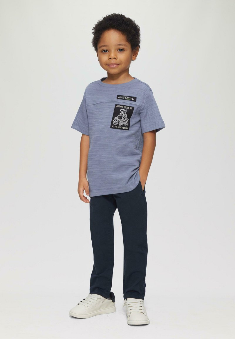 Grijze t-shirt met korte mouwen met een zwarte patch met witte graphics, gecombineerd met donkerblauwe broeken en witte sneakers met zwarte accenten.