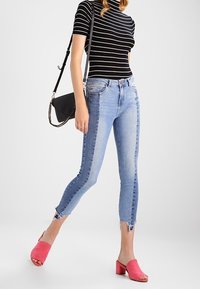 Úzké světle modré džíny se třepeným lemem; doplněné o černobílé pruhované tričko. Doplňky zahrnují malou černou kabelku a růžové mule boty.