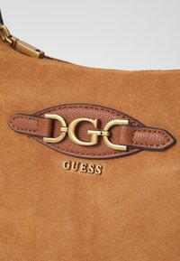 Sac à main en cuir suédé marron avec un logo et des accessoires en or. Présente des accents en cuir embossé et une texture lisse. Fermeture éclair sur le dessus.