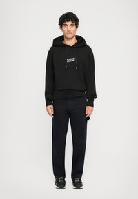 Čierna mikina s kapucňou a logom "VERSACE COUTURE", so šnúrkami, v kombinácii s tmavými džínsami a čiernymi športovými topánkami. Model stojí na jednofarebnom pozadí.