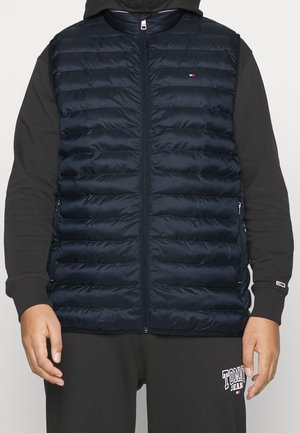 Man som bär en marinblå vadderad zip-up väst över en svart hoodie och svarta sweatpants med vit logotext på låret.