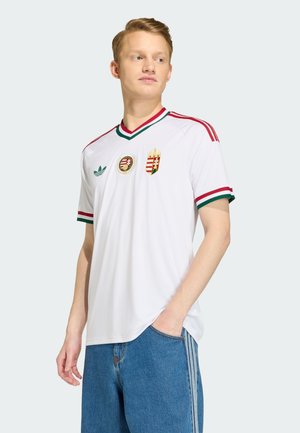 Junger Mann, der ein weißes Sporttrikot mit roten und grünen Abschlüssen und Ungarn-Wappen trägt, steht mit einer Hand in der Tasche und schaut zur Seite.