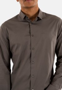 Camisa gris de botones con cuello, confeccionada en una tela texturizada, con un sutil logotipo y seis botones frontales. Mangas largas visibles en los puños.
