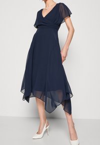 Marinblå chiffongklänning med V-ringning, korta volangärmar och asymmetrisk fåll. I kombination med vita högklackade skor.