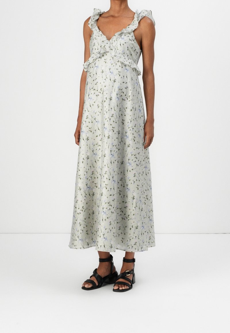 Femme portant une robe maxi sans manches légère à motifs floraux avec des bretelles volantées et des sandales noires à brides sur un fond uni.