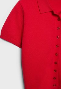 Chemise tricotée rouge à manches courtes avec col et une rangée verticale de sept boutons rouges assortis sur un fond clair.
