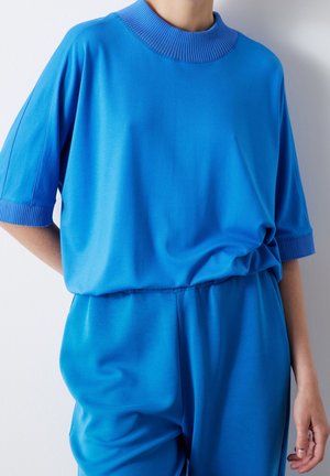 Blue jersey top with a ribbed collar, dropped shoulders, and loose fit, paired with matching blue pants, smooth texture, minimal details.