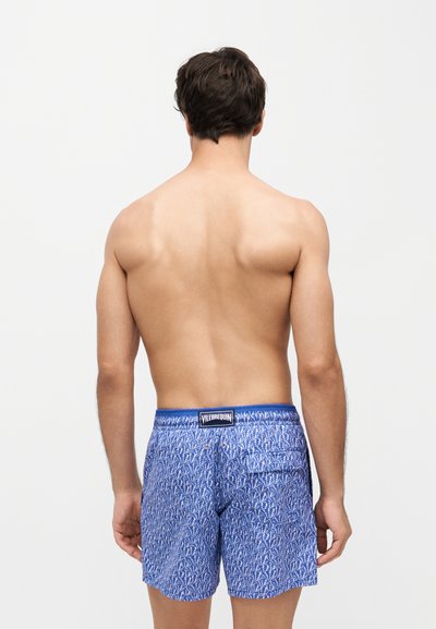 Homme debout, dos tourné vers l'avant, portant un short de bain bleu à motifs avec une poche arrière et une étiquette de marque à la taille.