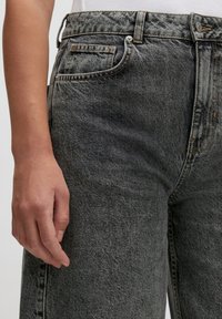 Hoge taille grijze denim jeans met een vervaagde textuur, voorzakken en contrasterende stiksels. Close-up van de taille en hand.