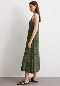 Grünes ärmelloses Maxikleid mit einem strukturierten, gepunkteten Muster. Kombiniert mit schwarzen Sandalen, die eine goldene Schnallenakzentuierung aufweisen.
