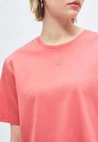 Sieviete valkā korāļrozā T-kreklu ar mazu apaļu logotipu centrā pie kakla. Attēls ir kadrēts pie zoda un pleciem.