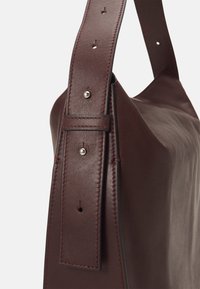 Bordeaux leren handtas met een gladde textuur, voorzien van een brede schouderband met klinknagels en zichtbare stiksels aan de zijkanten.