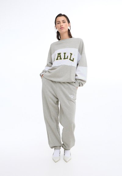 Felpa grigia con pannello bianco e scritta in grassetto "ALL". I pantaloni della tuta grigi abbinati presentano un logo. Il modello indossa scarpe bianche a punta.