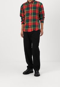 Červená, zelená a žlutá károvaná košile s dlouhými rukávy, kombinovaná s černými volnými kalhotami a černými členkovými botami.