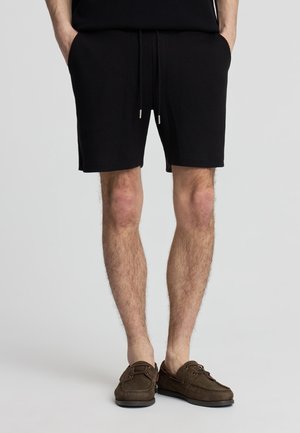 Uomo che indossa pantaloncini neri con coulisse e scarpe da barca in pelle scamosciata marrone, in piedi con le mani nelle tasche su uno sfondo semplice.