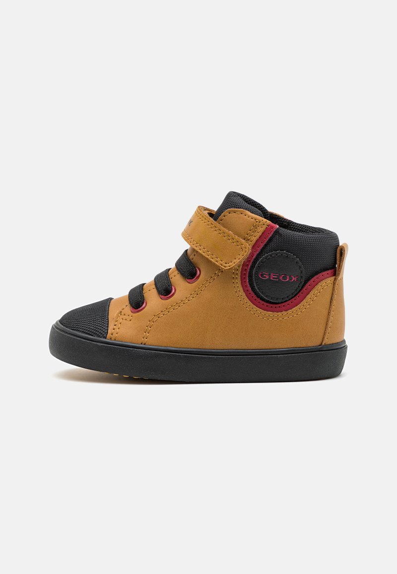 Zapato alto de cuero marrón con punta y suela de goma negra, acentos rojos, cordones negros y una correa de velcro en el costado.