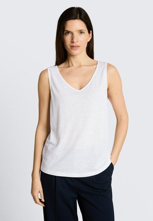 Femme aux longs cheveux bruns portant un top blanc sans manches à col en V et un pantalon foncé, debout avec une main dans la poche devant un fond uni.