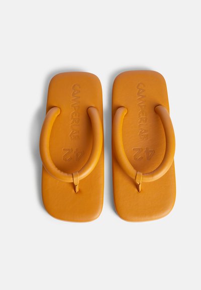 Oranssit kumiset tossut paksuilla, kaarevilla remmeillä ja tasaisella pohjalla. Yllä oleva pinta on varustettu kohotetulla logolla. Sileä pinta ja minimaalinen muotoilu.