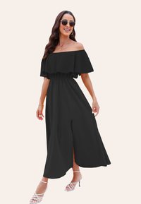 Maxi dress nera off-the-shoulder con maniche a volant, vita elastica e spacco alto; indossata con tacchi bianchi con cinturini e occhiali da sole.